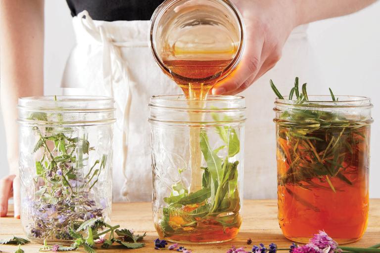 A woman in a white apron pouring honey into jars of herbs.